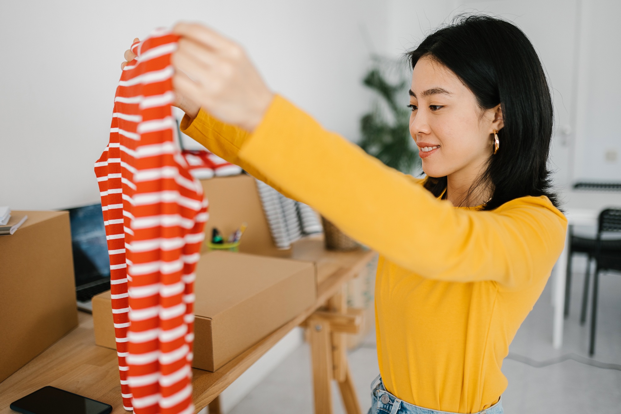 Smiling young asian woman unpacking clothes while sitting at desk
